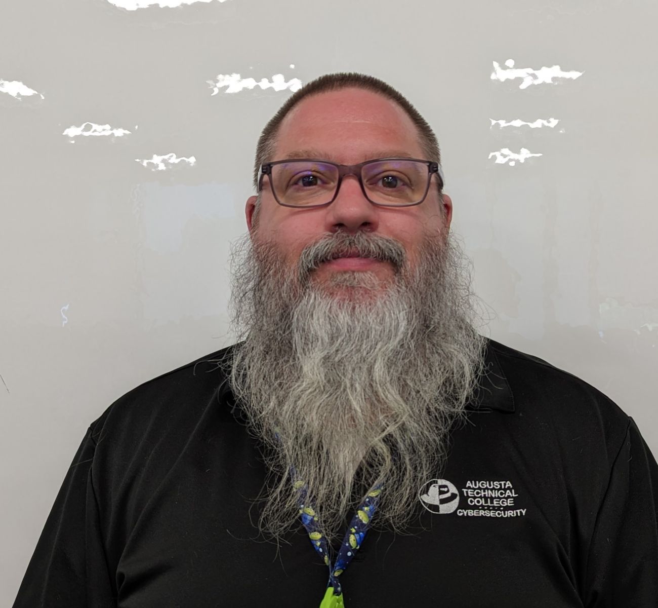 A Caucasian male with short brown hair and grey beard wearing a black Augusta Technical College Cybersecurity shirt. 